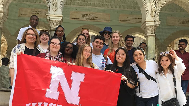 Students hoist a red Nebraska flag in front of an ornate colonnade.