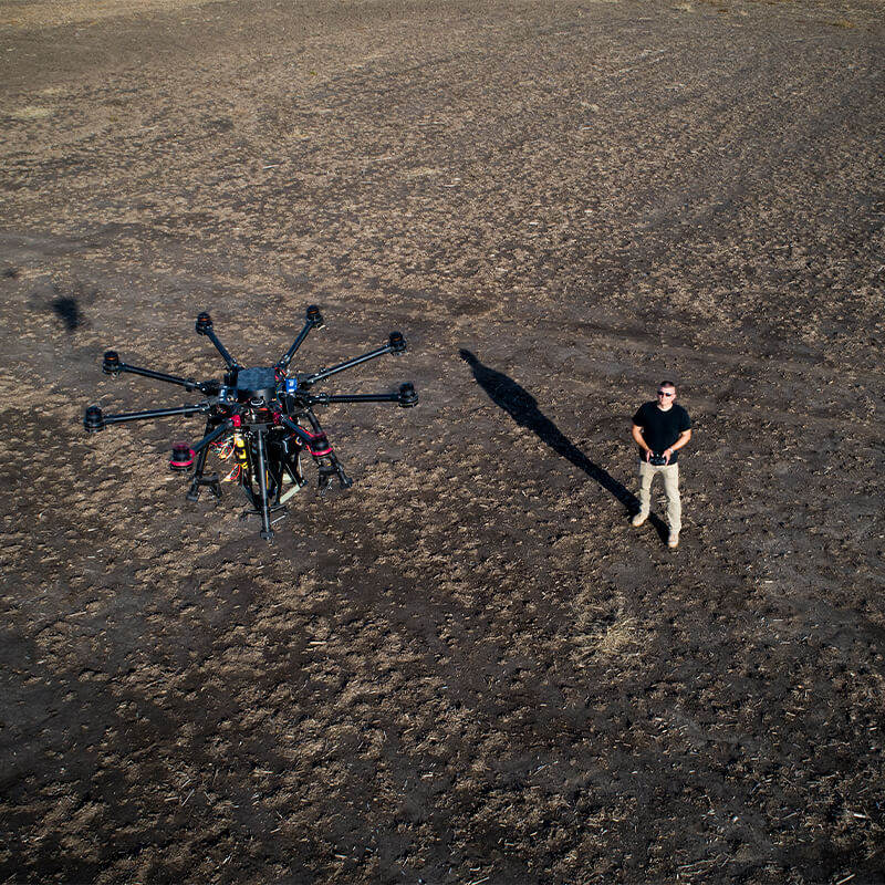Man in background piloting drone in foreground in barren field.