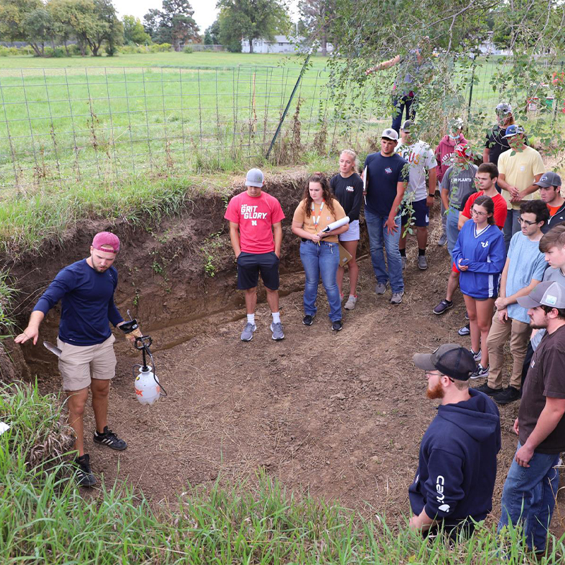Students in large pit watch and listen to instructor pointing at different soil strata.