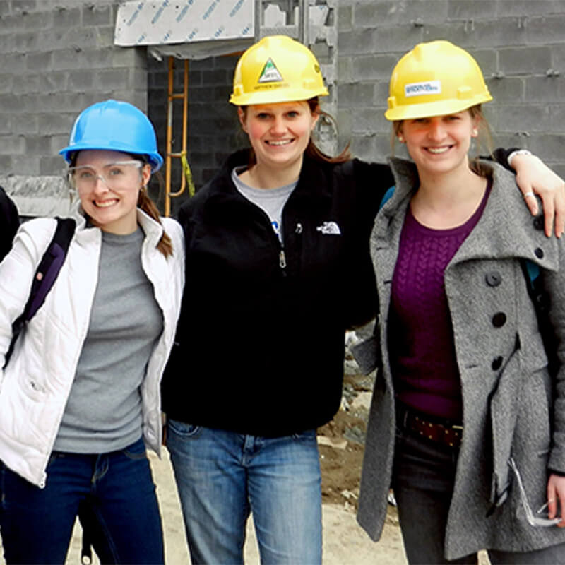 Three female students wearing hardhats smile at camera at construction site.