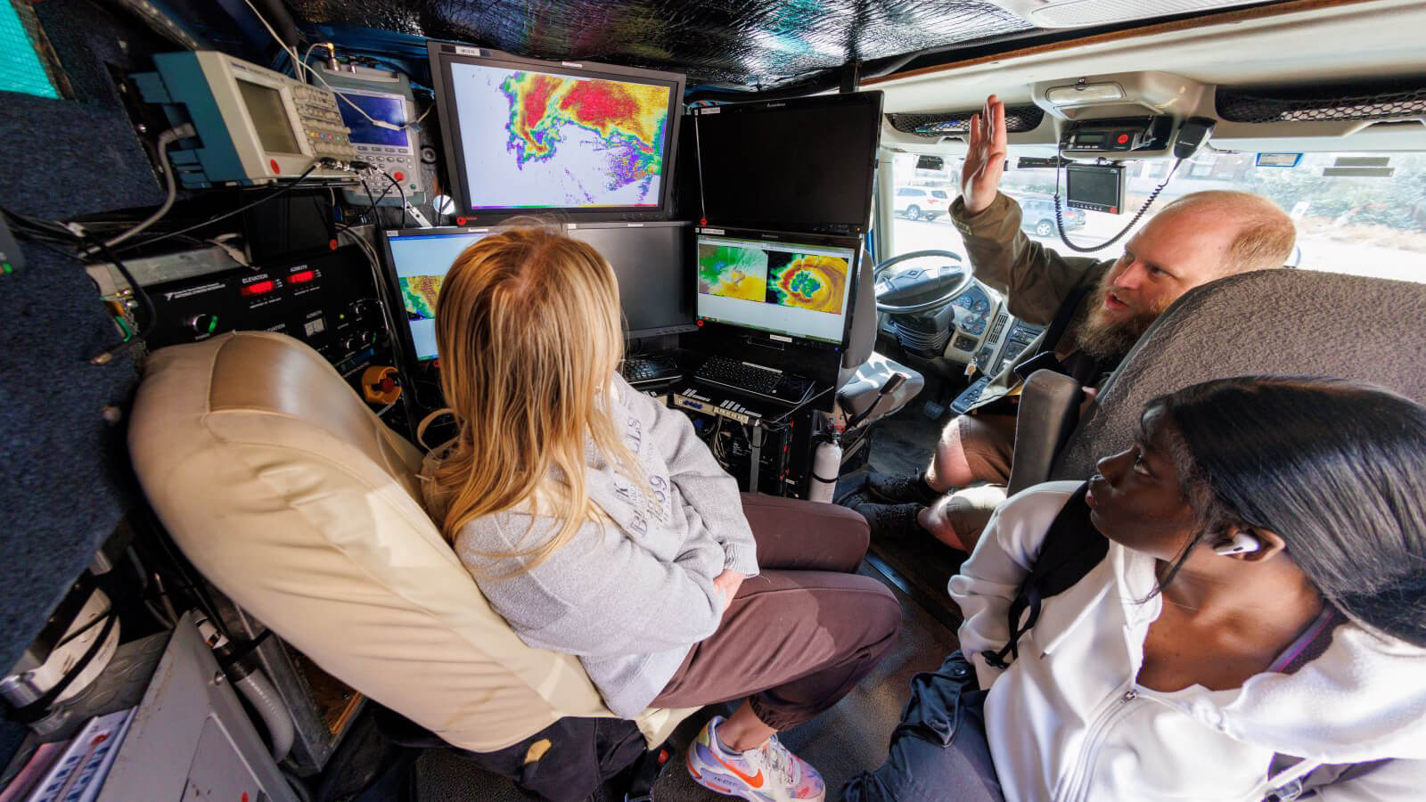 Students sit in storm chasing vehicle looking at screen filled with data with man explaining.
