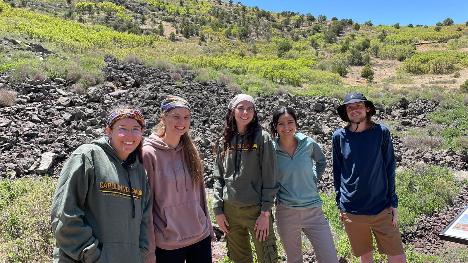 A group of students stands in front of a rock field on a rugged and arid looking hillside.