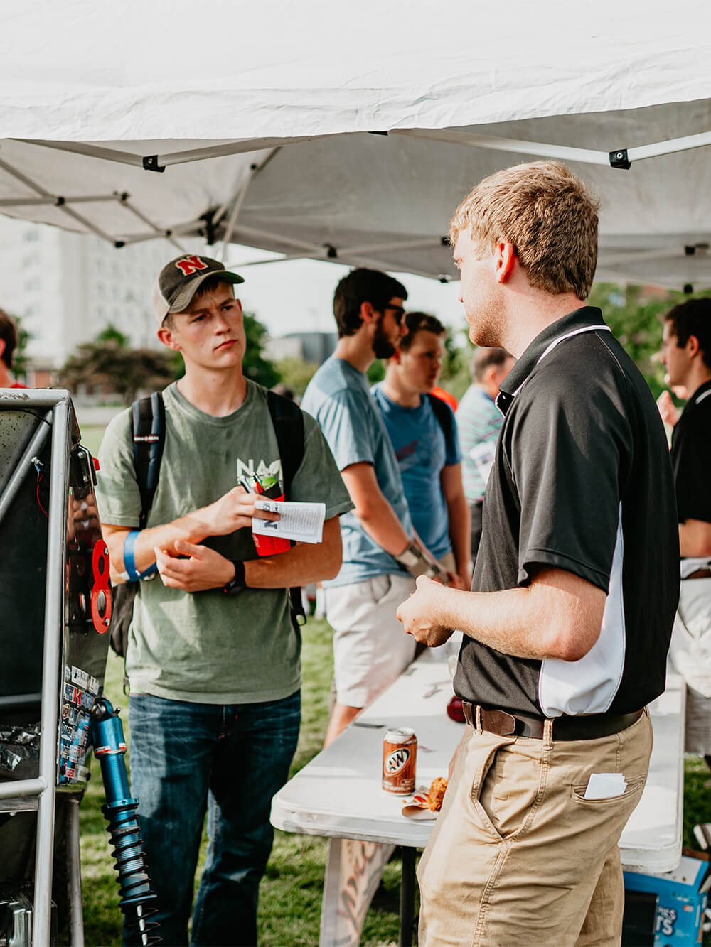 Student listens to man talking under sun tent during an outdoor event.