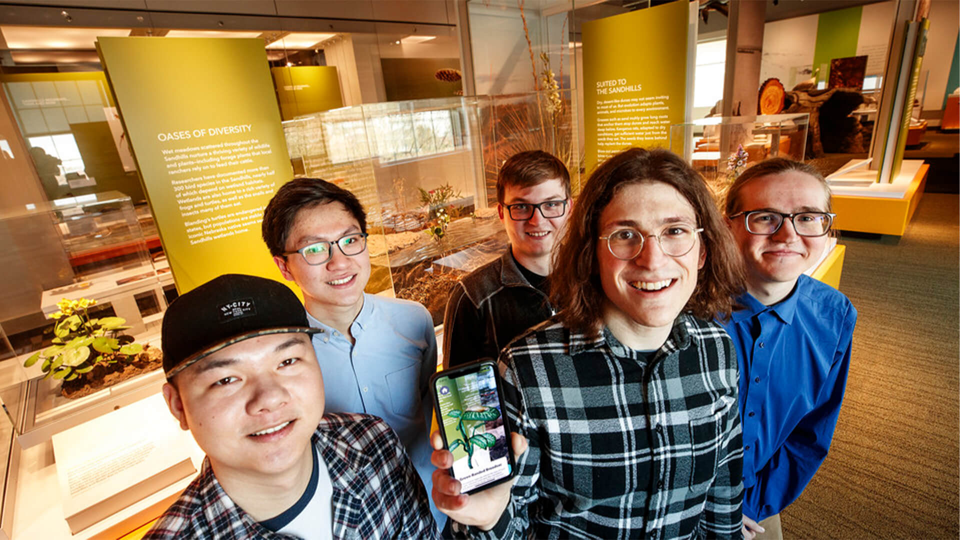 Group of students show off their app in front of Oasis of Diversity exhibition at museum.