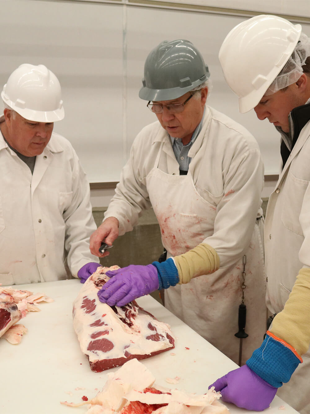 Three men in white butchers attire and hardhats cut large slab of meat