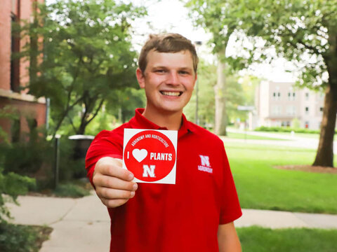 Man in red Agronomy polo shirt holds out a I HEART Plants sticker
