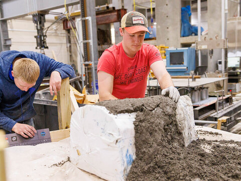 Student in red t-shirt put concrete over arch-like form while another student checks laptop.
