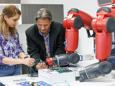 Student and professor work together in robotics lab with red robotic arms at right.