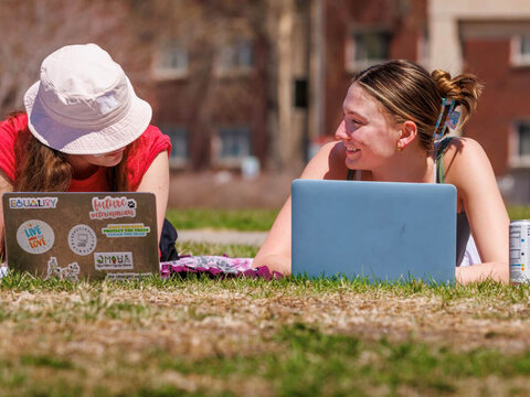 Students with laptops open talk outdoors on a sunny day.